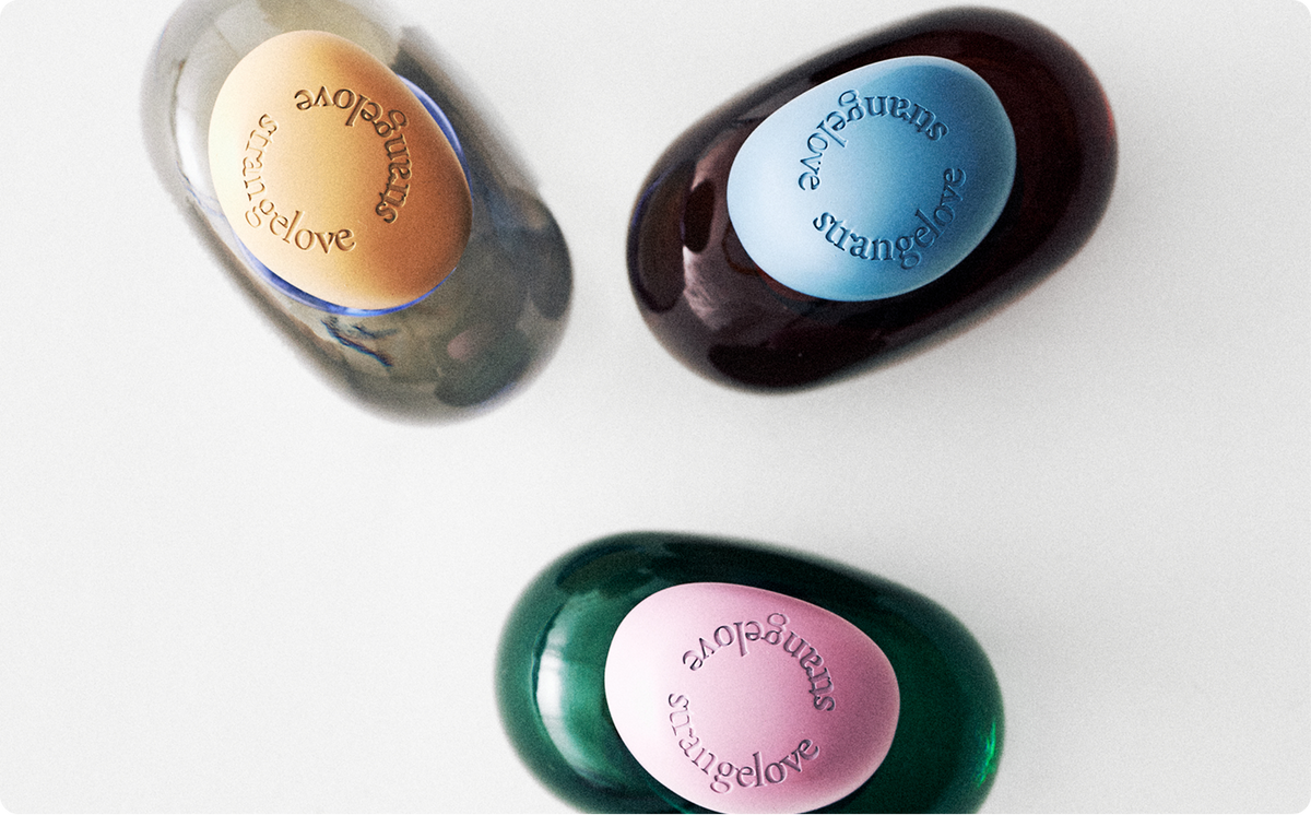 Three glossy oval soap bars shaped like oranges, labeled 'strangelove', in different colors: tan, dark brown, and emerald green, arranged on a white background.
