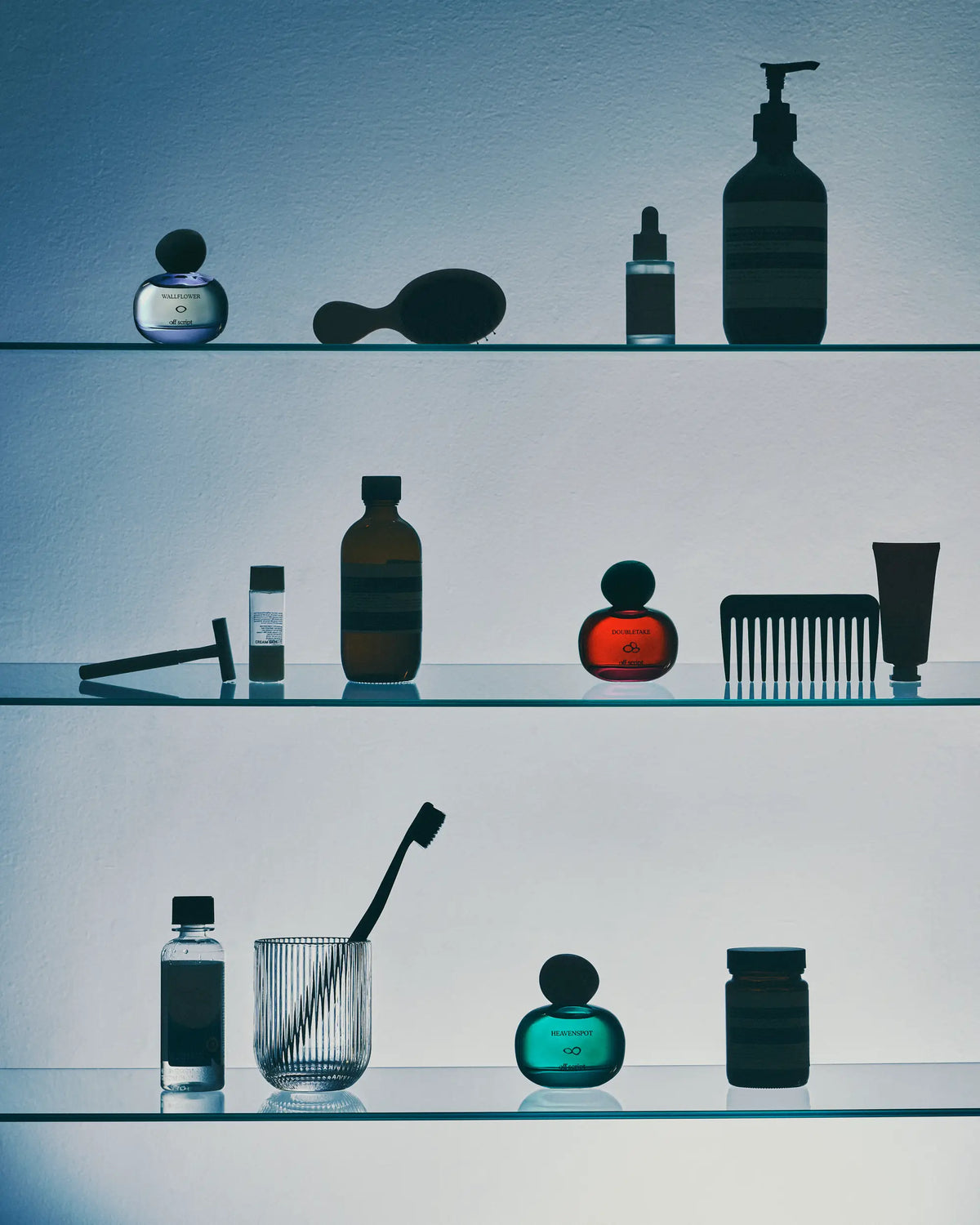 Three glass shelves displaying an assortment of toiletry and grooming products, including bottles, a comb, a brush, a toothbrush in a holder, and a small jar, set against a cool-toned blue-gray background.