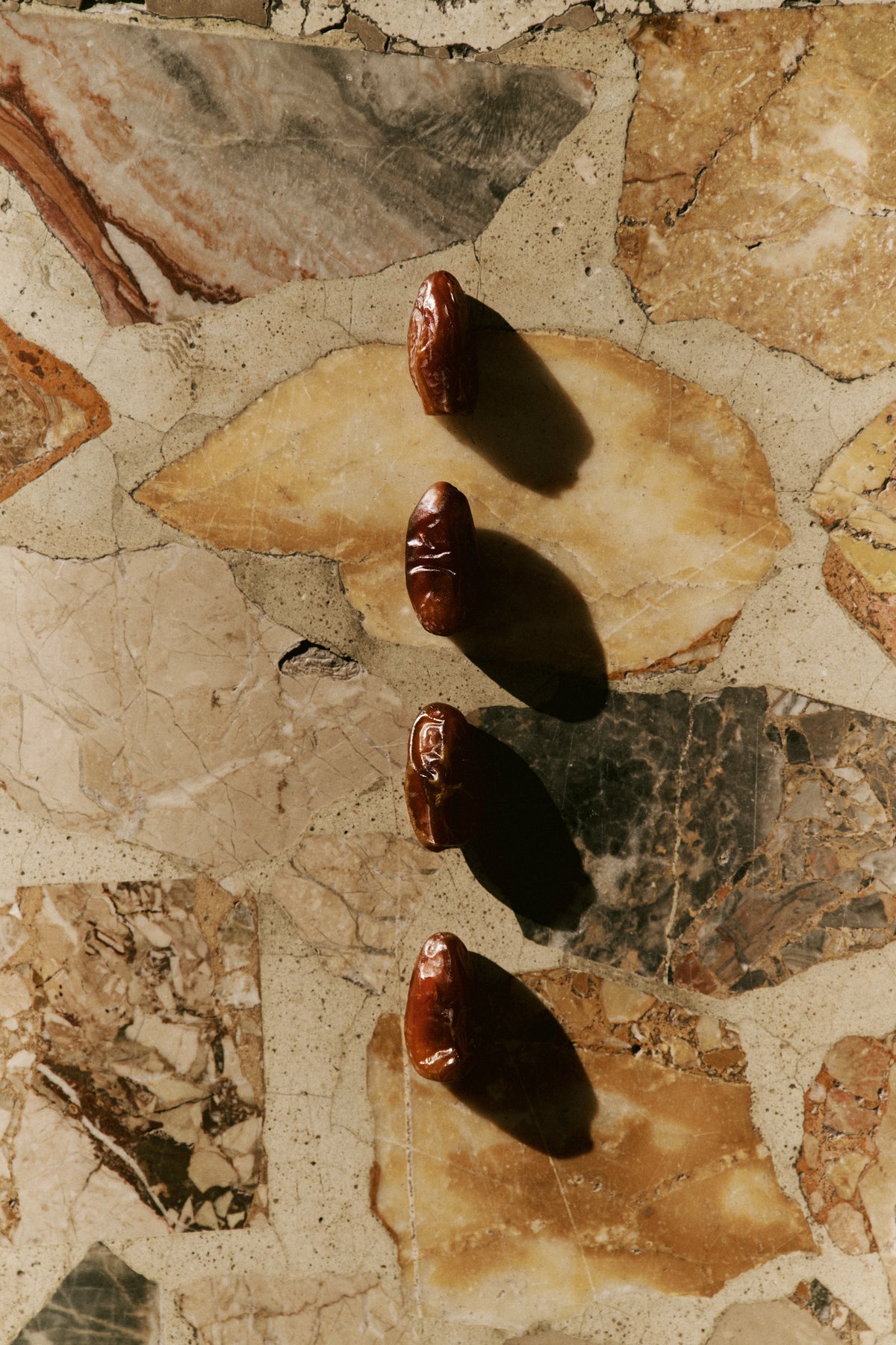 Four dark brown dates arranged vertically on a beige and brown stone tile surface, casting strong shadows under direct sunlight.