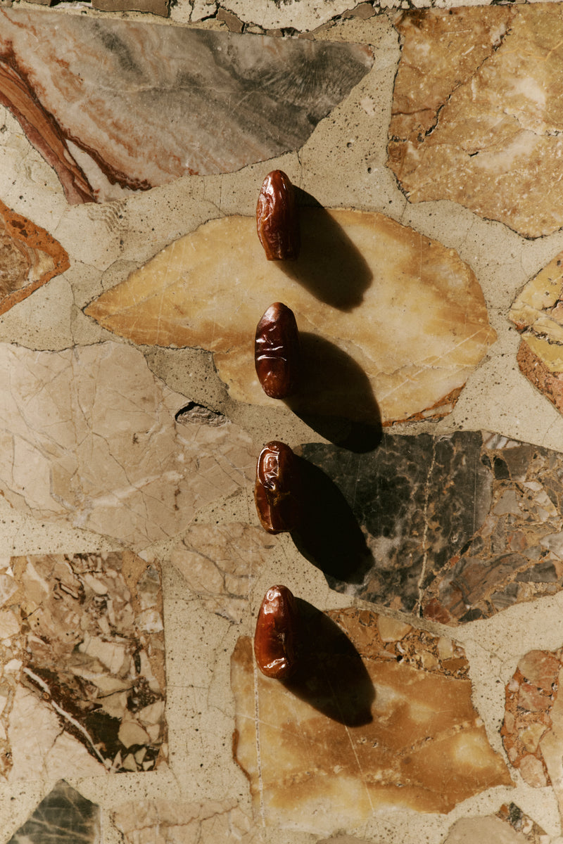 Four dark brown dates arranged vertically on a beige and brown stone tile surface, casting strong shadows under direct sunlight.