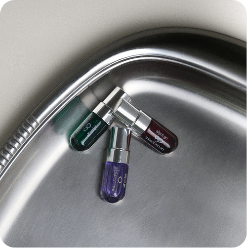 Three small cylindrical cosmetic tubes with metallic caps resting on the curved edge of a stainless steel sink. The tubes are dark green, dark brown, and purple, with visible branding for 'off script'.