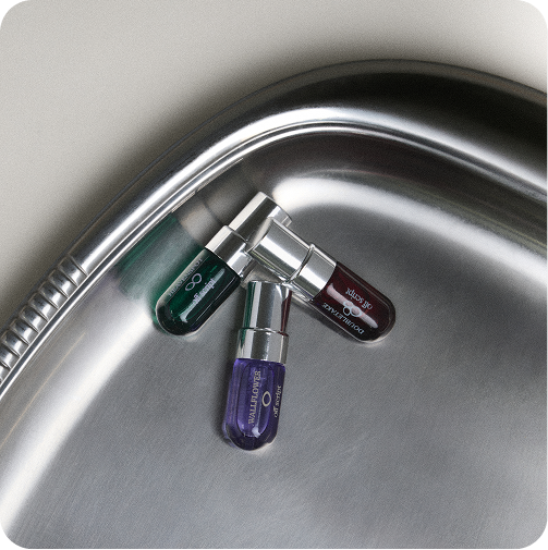 Three small cylindrical cosmetic tubes with metallic caps resting on the curved edge of a stainless steel sink. The tubes are dark green, dark brown, and purple, with visible branding for 'off script'.