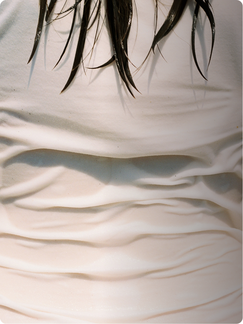 A close-up of a white or off-white fabric surface with soft folds and shadows, and a few dark hair strands visible at the top edge.