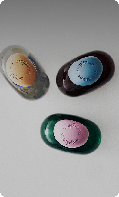 Three glossy oval soap bars shaped like oranges, labeled 'strangelove', in different colors: tan, dark brown, and emerald green, arranged on a white background.
